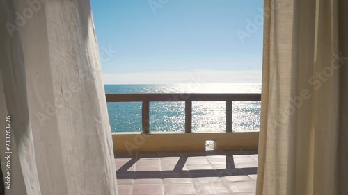 White curtains blow in slow motion on the balcony of an oceanfront resort hotel. Inside looking out at sunshine glimmering on the ocean surface. Peaceful shot embodies authentic travel experiences.