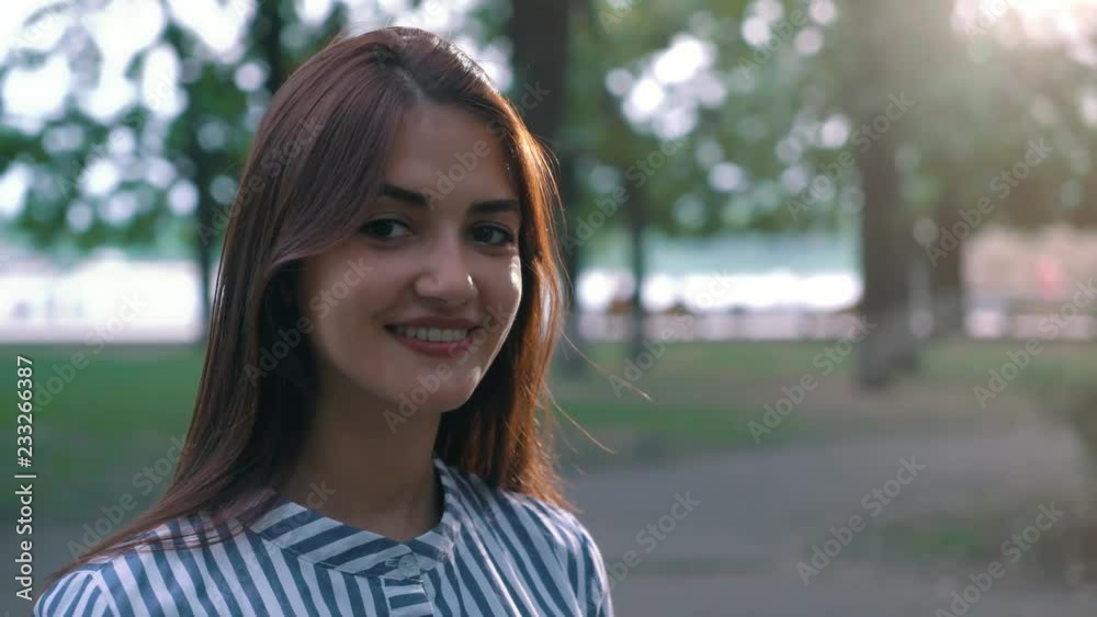 Portrait of a funny young woman with playful eyes and long loose hair in striped dress standing and smiling happily in a green park in summer. Portrait of a funny young woman with playful eyes and long loose hair in striped dress standing and smiling happily in a green park in summer.