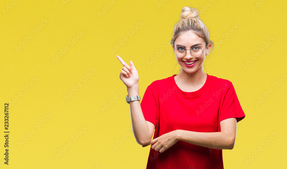 Young beautiful blonde woman wearing red t-shirt and glasses over isolated background with a big smile on face, pointing with hand and finger to the side looking at the camera.