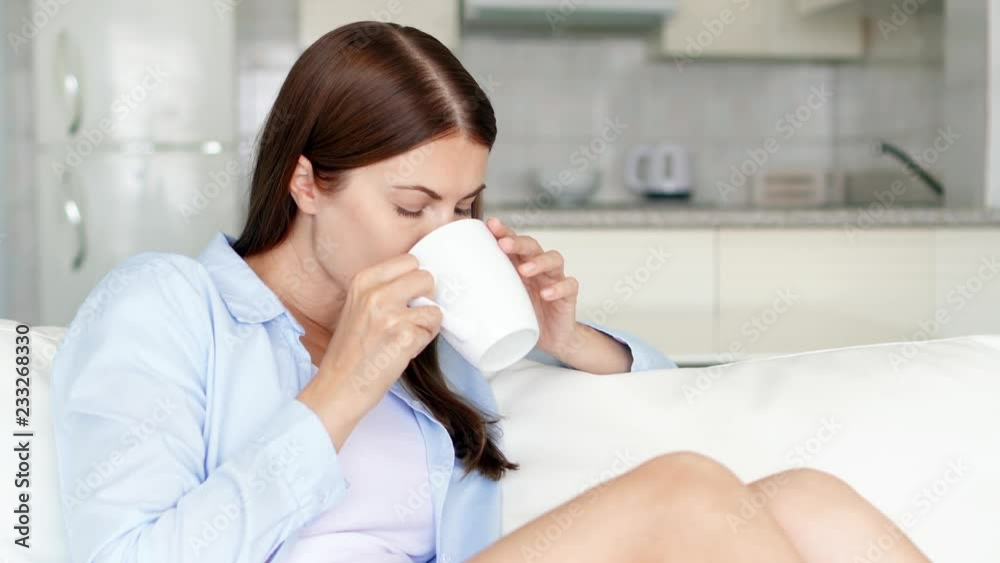 Young brunette woman with brown eyes relaxing sitting on couch at home in the morning. Pretty female housewife drinking tasty coffee or tea from white cup in living room. Kitchen on background Young brunette woman with brown eyes relaxing sitting on couch at home in the morning. Pretty female housewife drinking tasty coffee or tea from white cup in living room. Kitchen on background