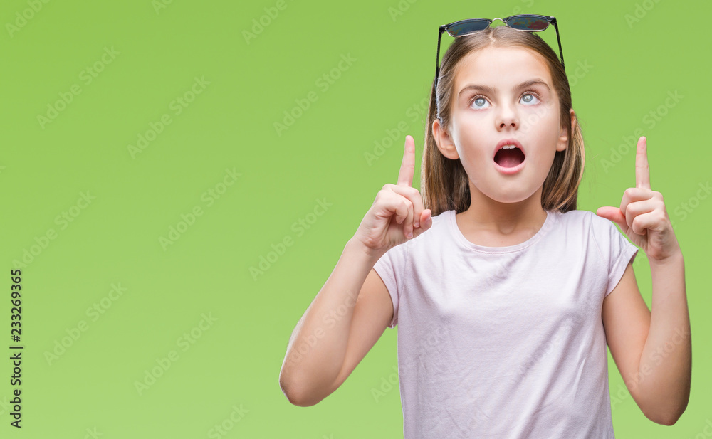 Young beautiful girl wearing sunglasses over isolated background amazed and surprised looking up and pointing with fingers and raised arms. Young beautiful girl wearing sunglasses over isolated background amazed and surprised looking up and pointing with fingers and raised arms.