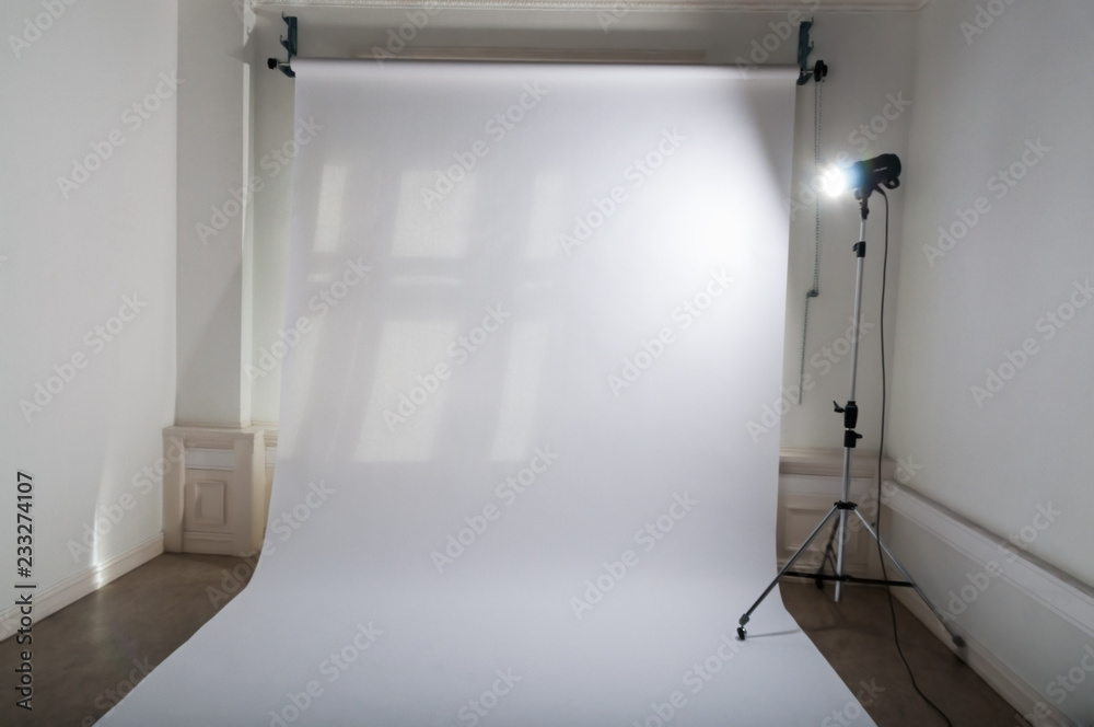 An empty professional photo studio with clean and simple photographic equipment setup of white, plain paper backdrop and a brightly lit flash spotlight on a lamp tripod in a dimmed atelier An empty professional photo studio with clean and simple photographic equipment setup of white, plain paper backdrop and a brightly lit flash spotlight on a lamp tripod in a dimmed atelier