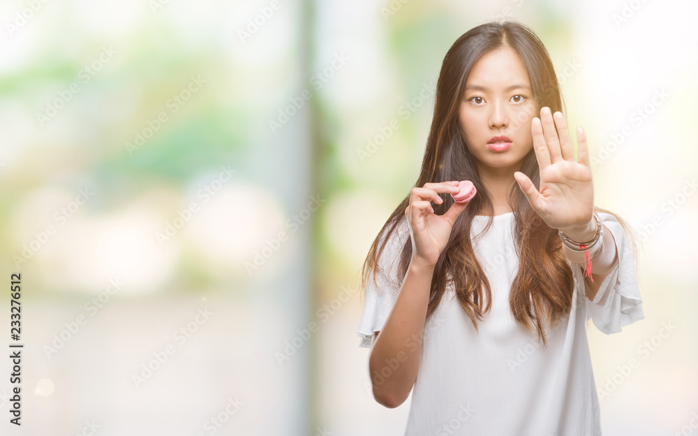 Young asian woman eating pink macaron sweet over isolated background with open hand doing stop sign with serious and confident expression, defense gesture Young asian woman eating pink macaron sweet over isolated background with open hand doing stop sign with serious and confident expression, defense gesture