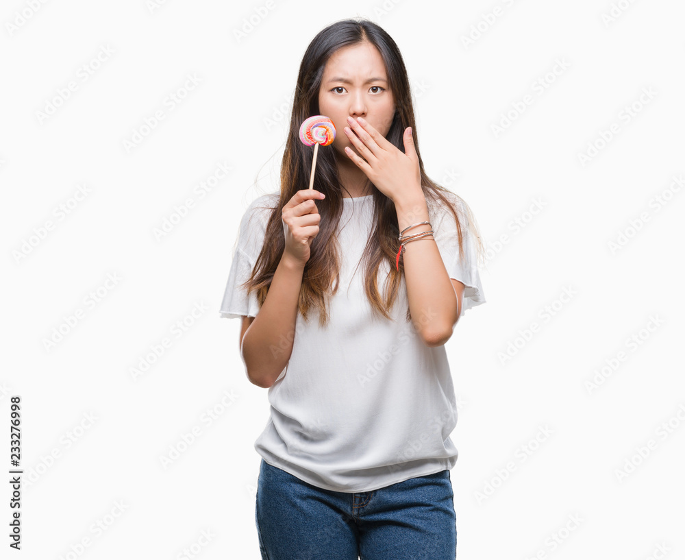 Young asian woman eating lollipop candy over isolated background cover mouth with hand shocked with shame for mistake, expression of fear, scared in silence, secret concept Young asian woman eating lollipop candy over isolated background cover mouth with hand shocked with shame for mistake, expression of fear, scared in silence, secret concept