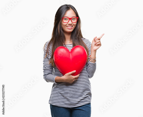 Young asian woman in love holding read heart over isolated background very happy pointing with hand and finger to the side