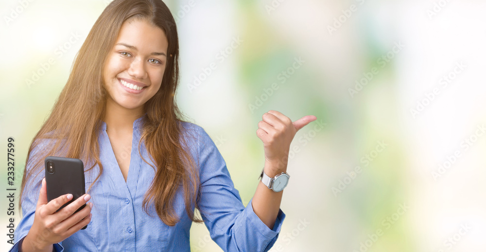 Young beautiful brunette business woman using smartphone over isolated background pointing and showing with thumb up to the side with happy face smiling
