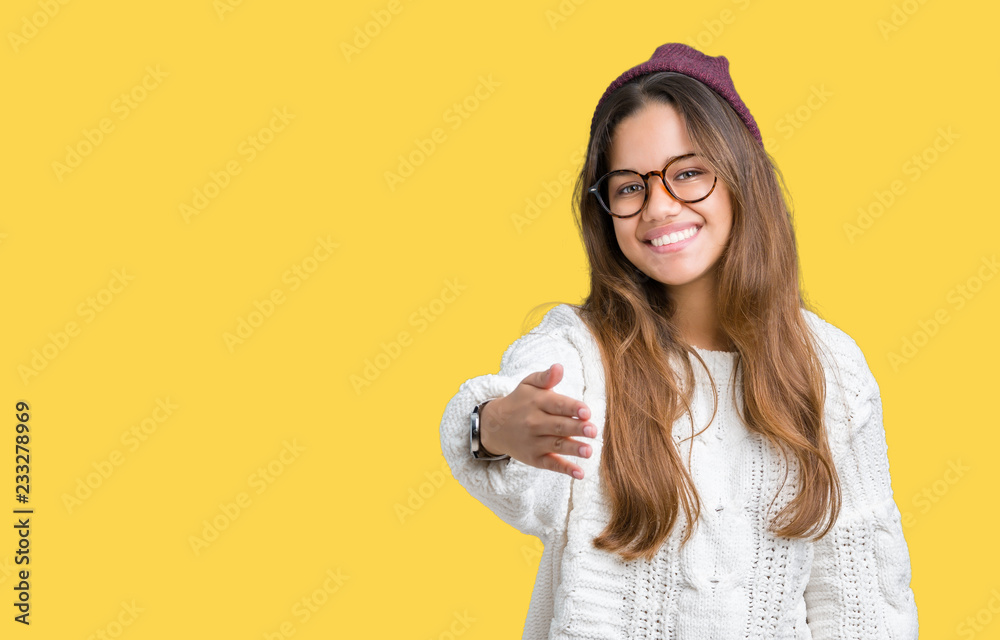Young beautiful brunette hipster woman wearing glasses and winter hat over isolated background smiling friendly offering handshake as greeting and welcoming. Successful business. Young beautiful brunette hipster woman wearing glasses and winter hat over isolated background smiling friendly offering handshake as greeting and welcoming. Successful business.