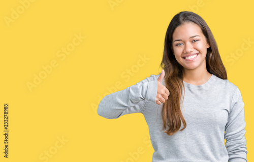 Young beautiful brunette woman wearing sweater over isolated background doing happy thumbs up gesture with hand. Approving expression looking at the camera with showing success.
