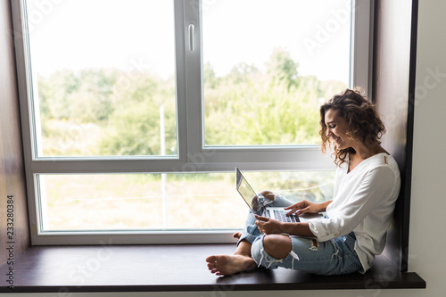 Beautiful young woman in white shirt, blue jeans sitting on windowsill laptop computer. Copy space, background. photo