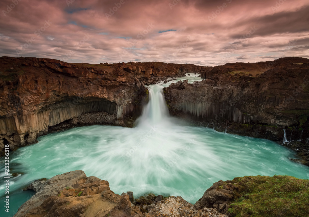 The waterfall Aldeyjarfoss with golden clouds in the sky. The flowing water is captured by a long exposure. Amazing blue color of water from the glacier. Natural and colorful environment. The waterfall Aldeyjarfoss with golden clouds in the sky. The flowing water is captured by a long exposure. Amazing blue color of water from the glacier. Natural and colorful environment.