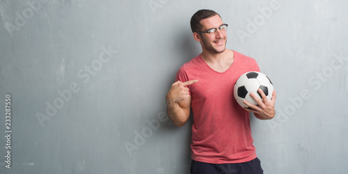 Young caucasian man over grey grunge wall holding soccer football ball very happy pointing with hand and finger