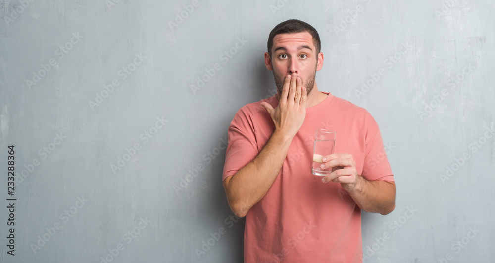 Young caucasian man over grey grunge wall drinking a glass of water cover mouth with hand shocked with shame for mistake, expression of fear, scared in silence, secret concept Young caucasian man over grey grunge wall drinking a glass of water cover mouth with hand shocked with shame for mistake, expression of fear, scared in silence, secret concept