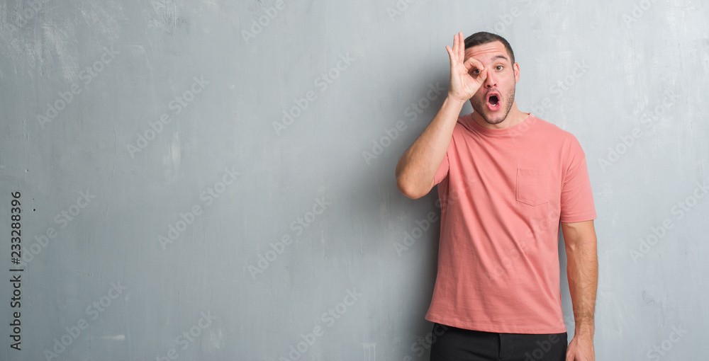 Young caucasian man over grey grunge wall doing ok gesture shocked with surprised face, eye looking through fingers. Unbelieving expression.