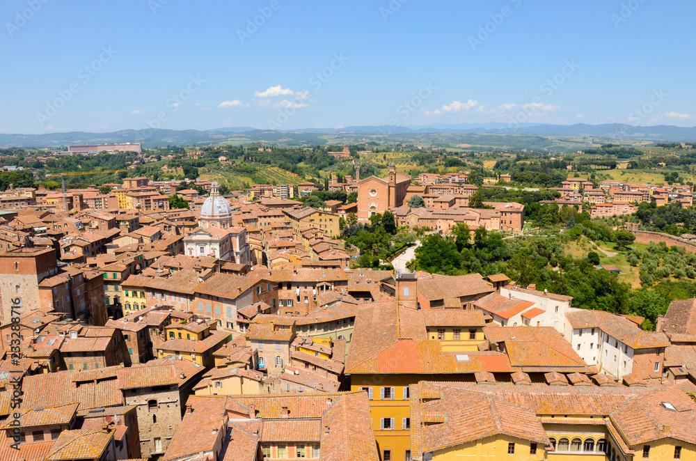 Rooftop view of colorful yellow, green, and brown buildings in the medieval city of Siena, Italy with the countryside of Tuscany in the distance under a beautiful blue sky
