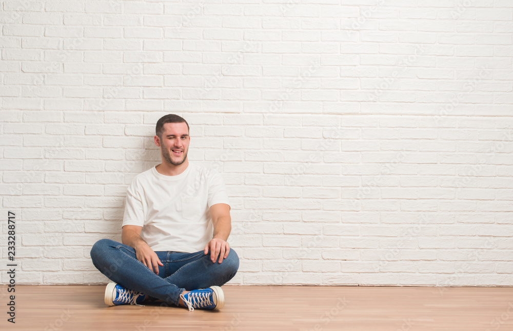 Young caucasian man sitting on the floor over white brick wall winking looking at the camera with sexy expression, cheerful and happy face. Young caucasian man sitting on the floor over white brick wall winking looking at the camera with sexy expression, cheerful and happy face.