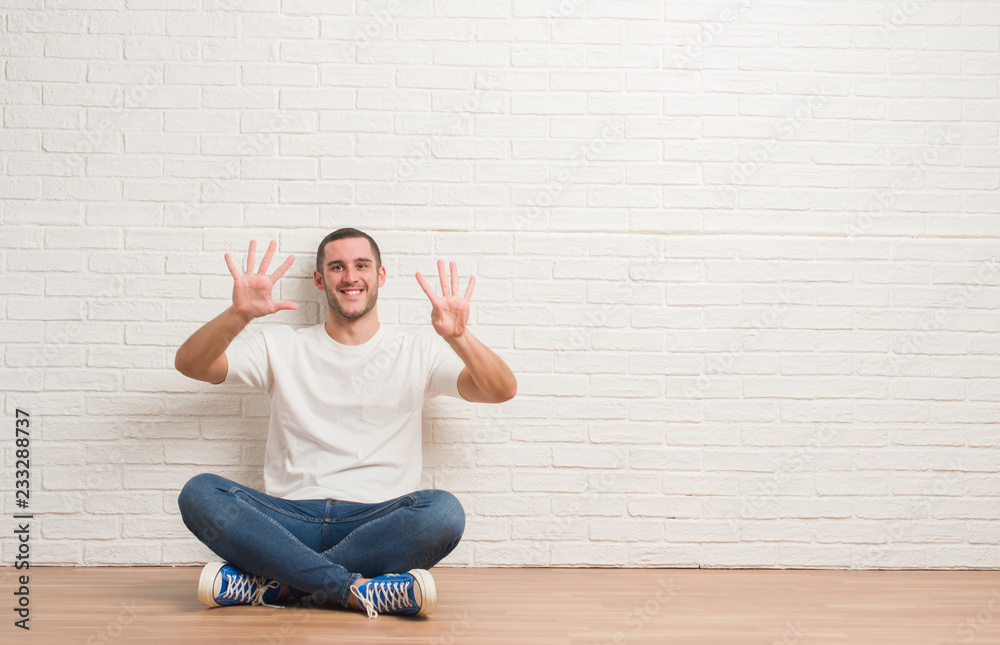 Young caucasian man sitting on the floor over white brick wall showing and pointing up with fingers number nine while smiling confident and happy. Young caucasian man sitting on the floor over white brick wall showing and pointing up with fingers number nine while smiling confident and happy.