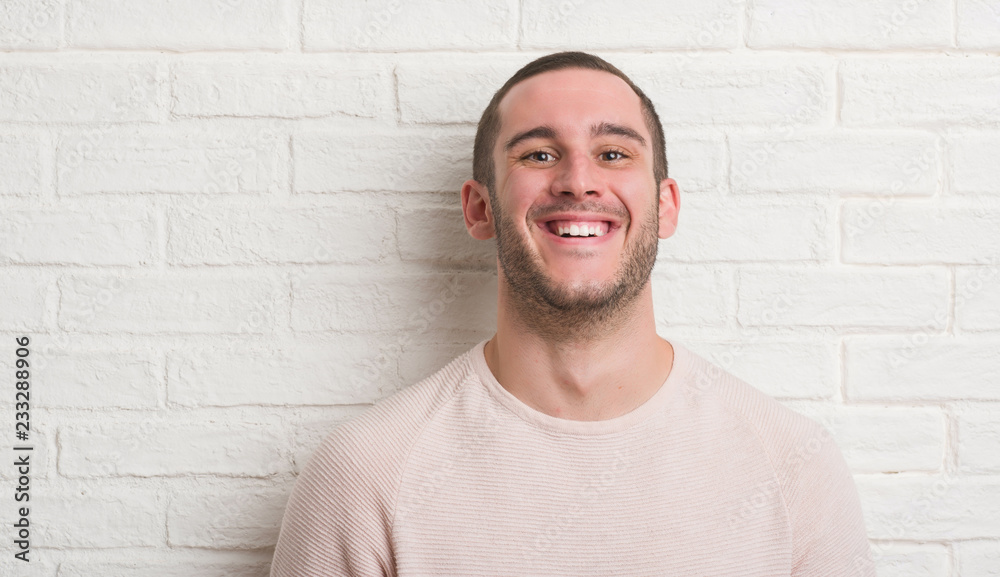Young caucasian man standing over white brick wall with a happy face standing and smiling with a confident smile showing teeth Young caucasian man standing over white brick wall with a happy face standing and smiling with a confident smile showing teeth