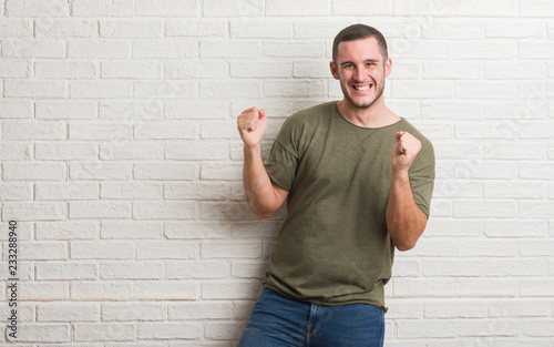 Young caucasian man standing over white brick wall very happy and excited doing winner gesture with arms raised, smiling and screaming for success. Celebration concept.