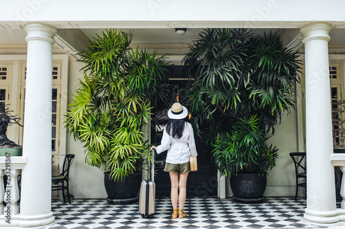 Back view of a woman on a vacation standing in front of a hotel and holding a suitcase.