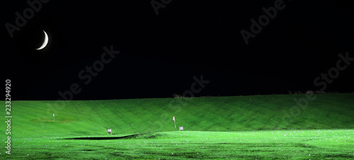 Illuminated golf driving range at night with flag and moon in the sky abive.