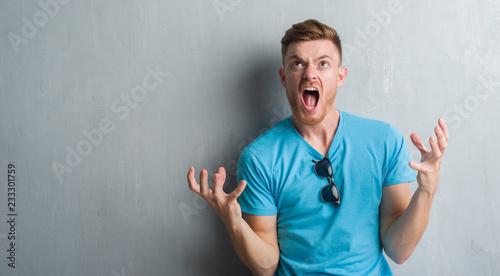 Young redhead man over grey grunge wall wearing casual outfit crazy and mad shouting and yelling with aggressive expression and arms raised. Frustration concept.