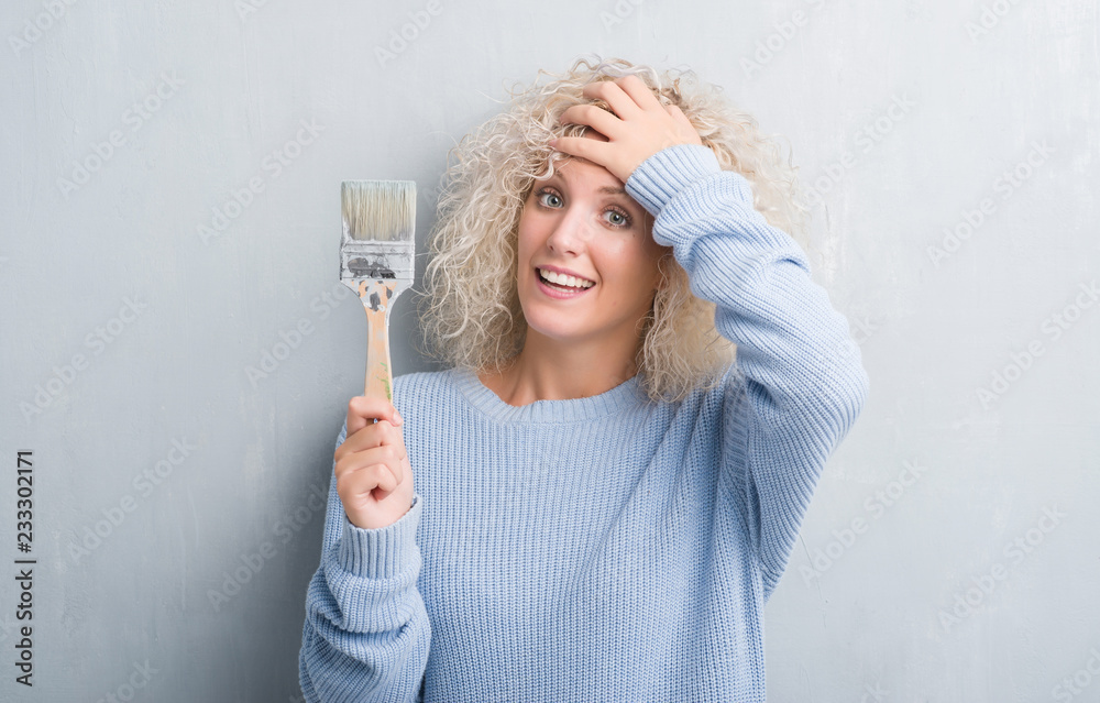 Young blonde woman with curly hair over grunge grey background holding paint brush stressed with hand on head, shocked with shame and surprise face, angry and frustrated. Fear and upset for mistake. Young blonde woman with curly hair over grunge grey background holding paint brush stressed with hand on head, shocked with shame and surprise face, angry and frustrated. Fear and upset for mistake.