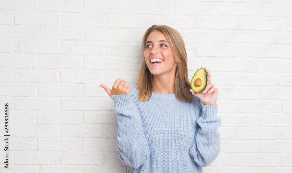 Beautiful young woman over white brick wall eating fresh avocado pointing and showing with thumb up to the side with happy face smiling Beautiful young woman over white brick wall eating fresh avocado pointing and showing with thumb up to the side with happy face smiling