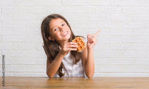Young hispanic kid sitting on the table eating waffle very happy pointing with hand and finger to the side