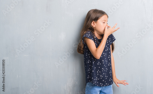 Young hispanic kid over grunge grey wall smelling something stinky and disgusting, intolerable smell, holding breath with fingers on nose. Bad smells concept.