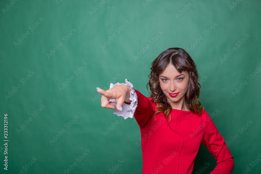 Image of cheerful woman with long brown hair winking and showing index finger aside meaning hey you isolated over green background.Emotional expressing woman in red dress, red lips and dark curly