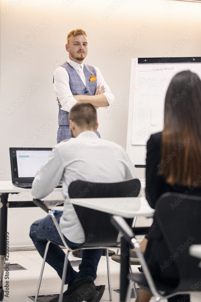 one young man, 20-29 years old, presenting to group of people, arms crossed, serious expression, in office indoors. one young man, 20-29 years old, presenting to group of people, arms crossed, serious expression, in office indoors.