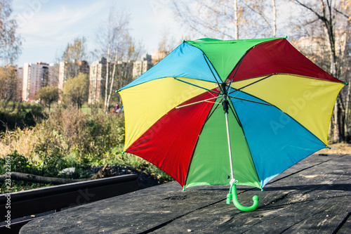 Fototapeta Naklejka Na Ścianę i Meble -  A multi-colored umbrella in red, green, yellow, blue is standing on a wooden table in the autumn park in the background are trees, new buildings and a pond. The concept of autumn, rainy autumn day
