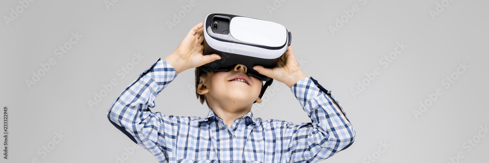A charming boy with an inlaid shirt and light jeans stands on a gray background. The boy on his face glasses virtual reality