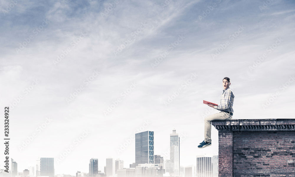 Man on roof edge reading book and cityscape at background Man on roof edge reading book and cityscape at background