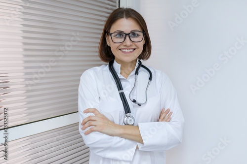 Portrait of positive confident adult female doctor with arms crossed near the door inside the hospital