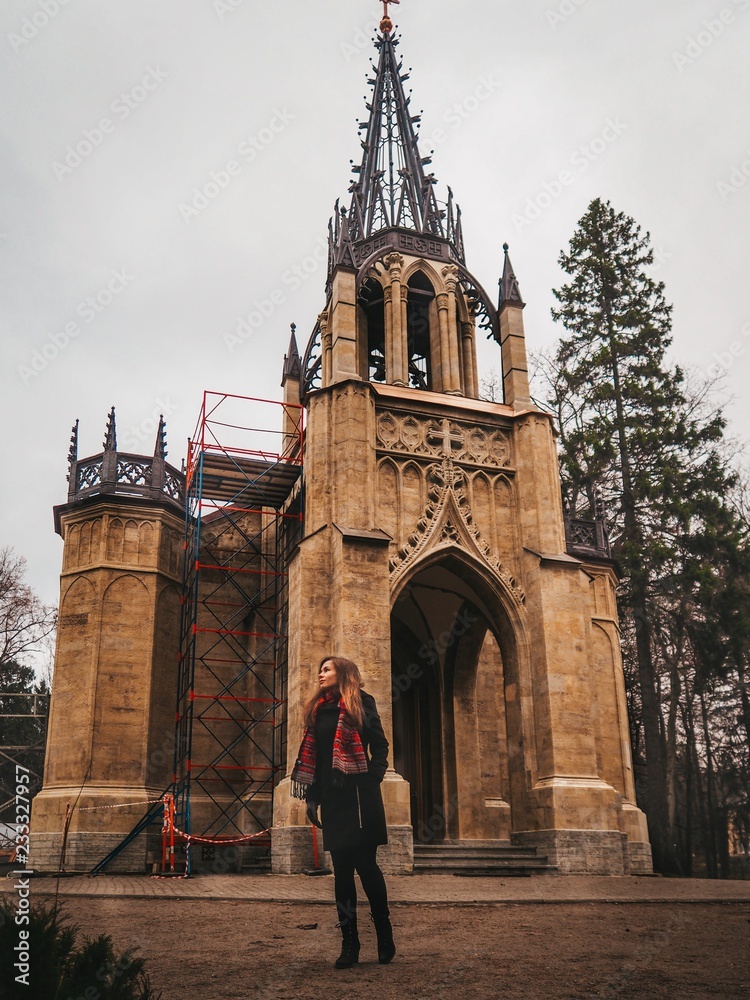 Brunette girl with long hair in black coat and bright red scarf stands near old Gothic Church / Cathedral in the forest