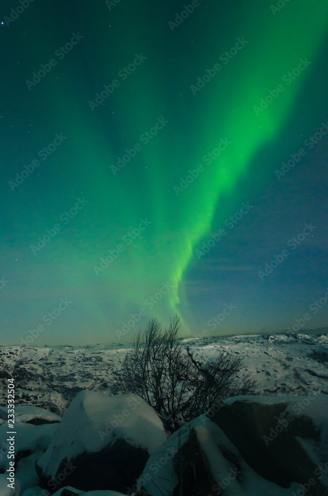 Beautiful northern lights, aurora in the night sky above the snow-covered hills. Large stones and a frozen lake.