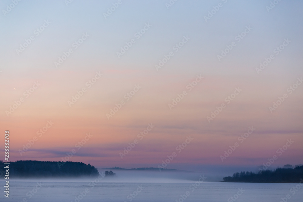 Christmas winter landscape of the frozen lake covered with snow on a cold winter day and the forest in thr mist 