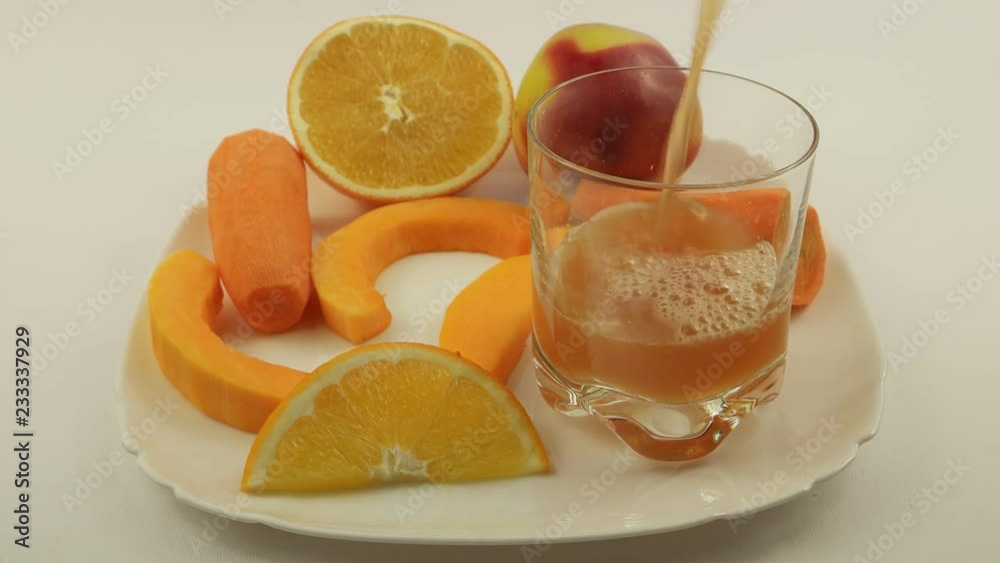 Transparent glass tumbler is filling with apple juice on the background of white dish with fruits and vegetables Transparent glass tumbler is filling with apple juice on the background of white dish with fruits and vegetables