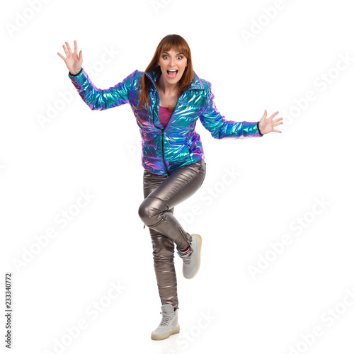 Young Woman In Vibrant Down Jacket Is Standing On One Leg With Arms Outstretched And Shouting