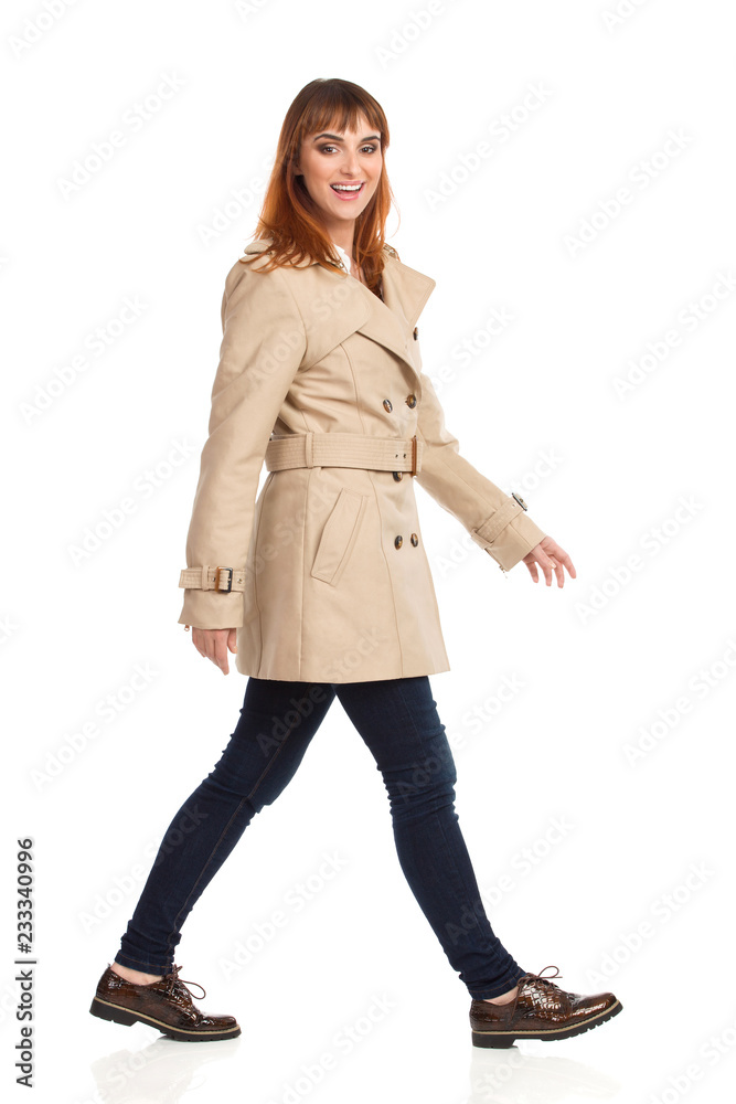 Young Woman In Beige Coat, Jeans And Brown Shoes Is Walking And Looking At Camera