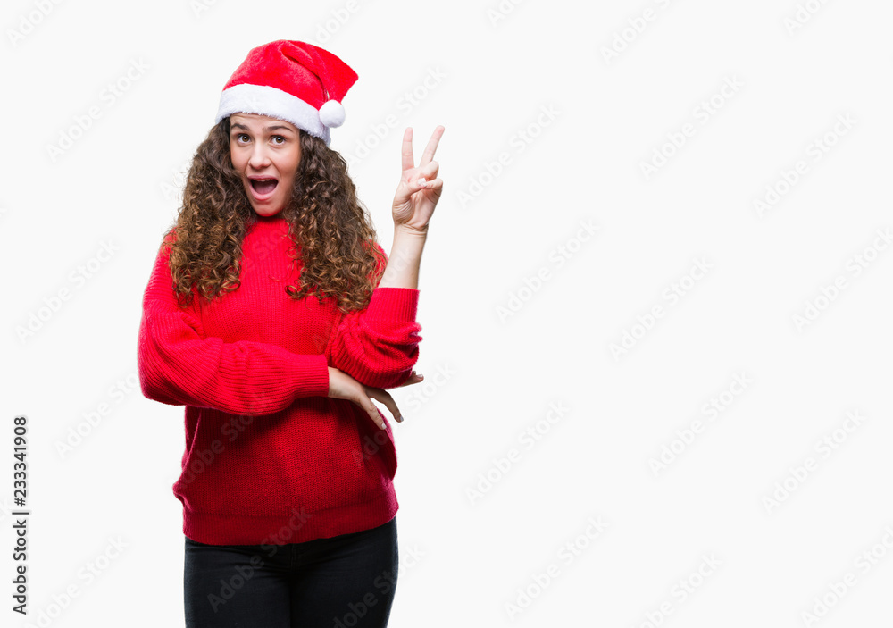 Young brunette girl wearing christmas hat over isolated background smiling with happy face winking at the camera doing victory sign. Number two. Young brunette girl wearing christmas hat over isolated background smiling with happy face winking at the camera doing victory sign. Number two.