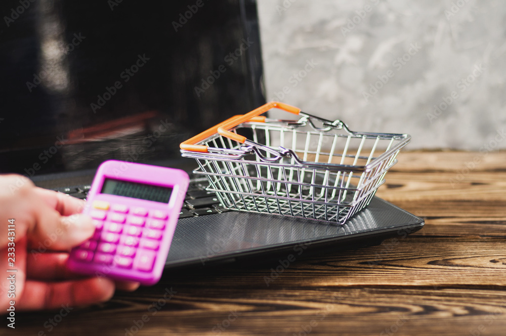 Man's hand holds calculator on background of old dirty laptop and empty market basket on brown wooden table. Online trade concept
