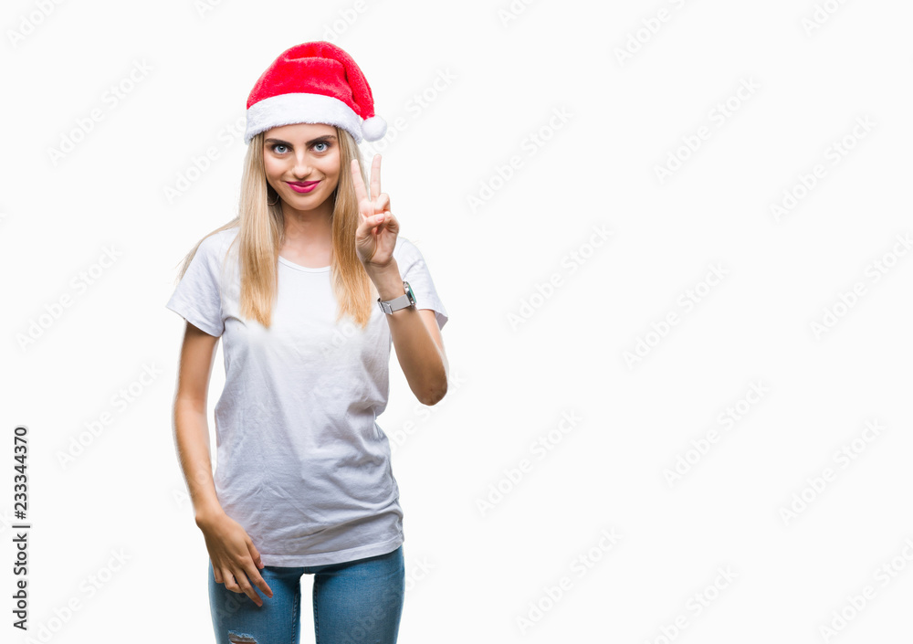 Young beautiful blonde woman christmas hat over isolated background smiling with happy face winking at the camera doing victory sign. Number two.