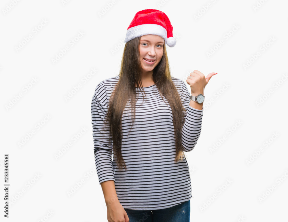 Young beautiful caucasian woman wearing christmas hat over isolated background smiling with happy face looking and pointing to the side with thumb up. Young beautiful caucasian woman wearing christmas hat over isolated background smiling with happy face looking and pointing to the side with thumb up.