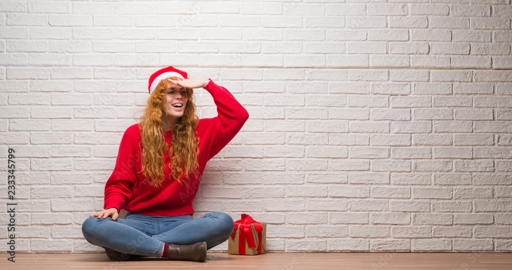 Young redhead woman sitting over brick wall wearing christmas hat very happy and smiling looking far away with hand over head. Searching concept. Young redhead woman sitting over brick wall wearing christmas hat very happy and smiling looking far away with hand over head. Searching concept.