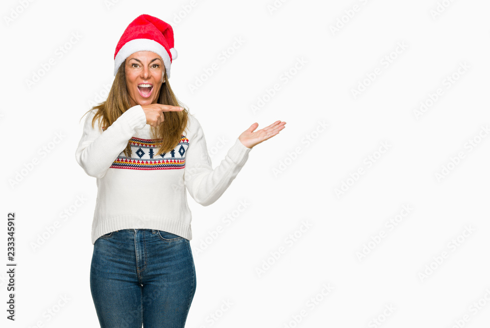 Middle age adult woman wearing winter sweater and chrismat hat over isolated background amazed and smiling to the camera while presenting with hand and pointing with finger. Middle age adult woman wearing winter sweater and chrismat hat over isolated background amazed and smiling to the camera while presenting with hand and pointing with finger.