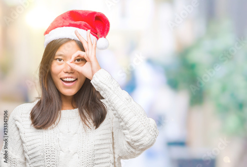 Young asian woman wearing christmas hat over isolated background doing ok gesture with hand smiling, eye looking through fingers with happy face.