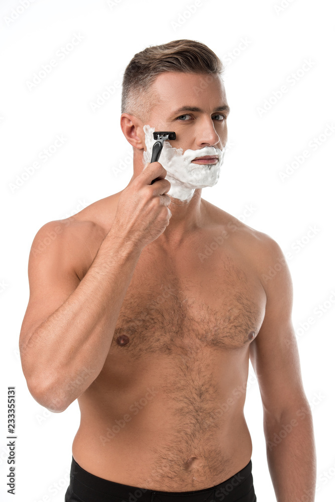 serious muscular shirtless man with foam on face shaving with razor isolated on white