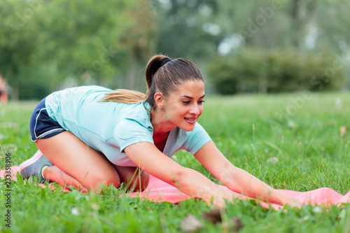 Smiling fit young woman practicing yoga in a park, stretching on an exercise mat. Active sport lifestyle in urban environment.